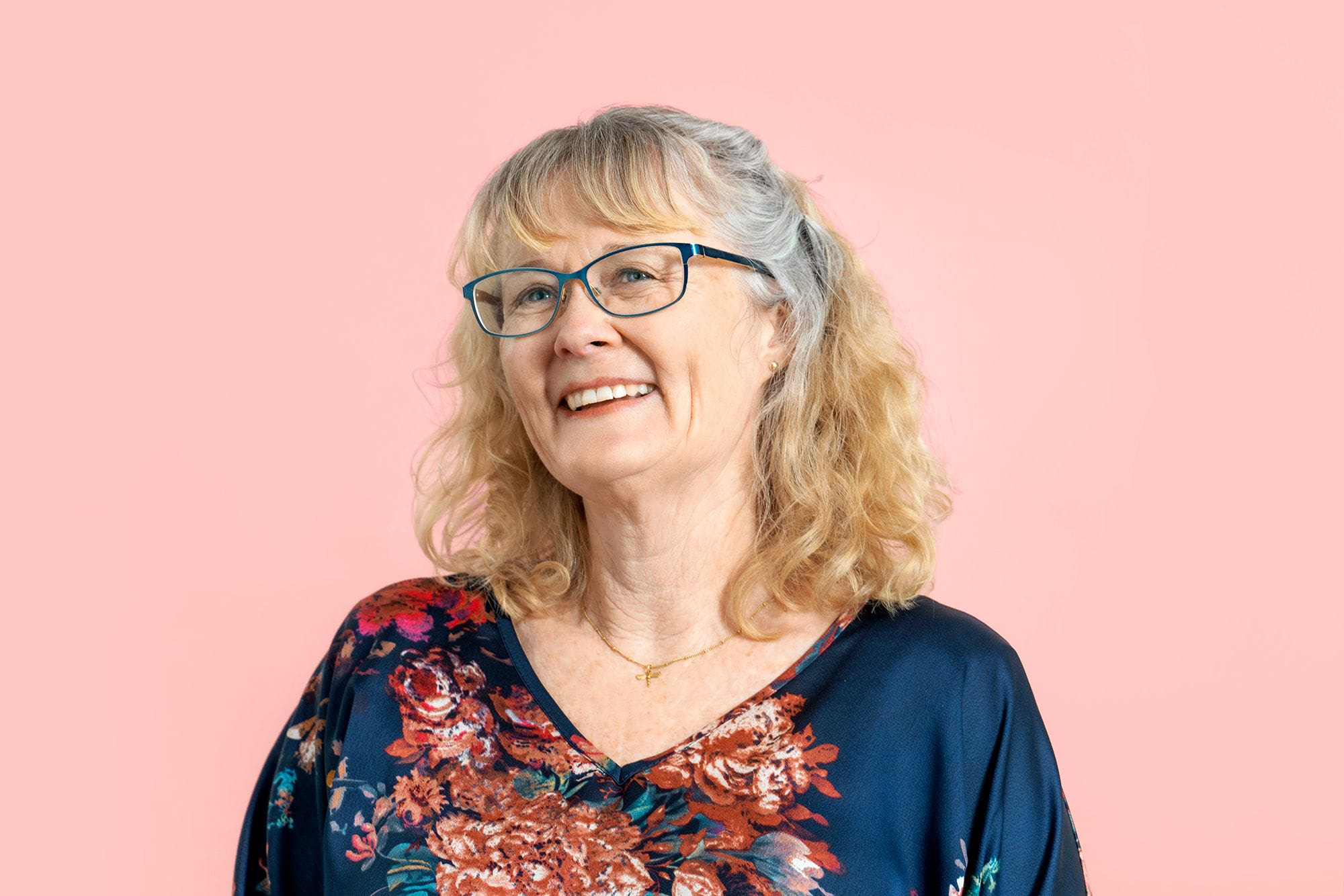 A colour photograph of Tracey, who has shoulder length blonde hair and blue eyes. She wears glasses. She smiles, standing in front of a light pink background.