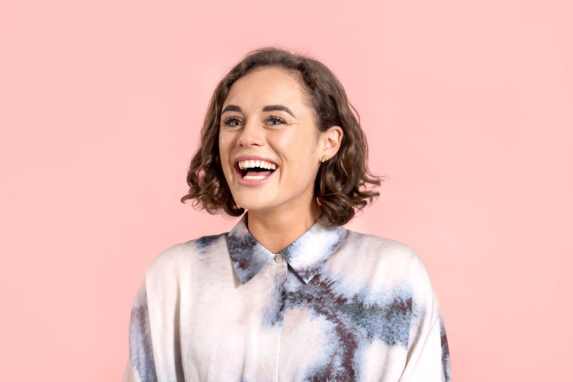 Sophie Jones has brown curly hair and green eyes. She laughs as she stands in front of a light pink background.