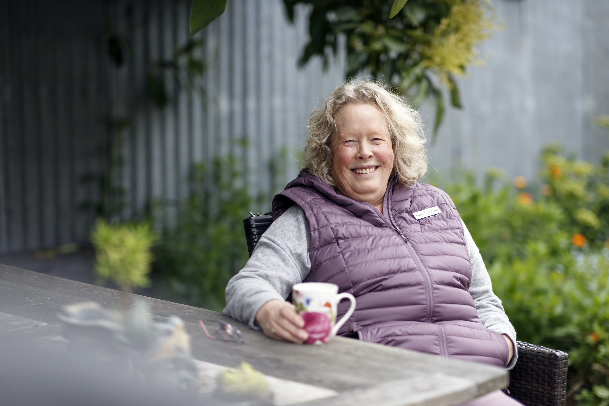 Karen holds a cup of tea as she sits in her garden. She smiles, wearing a gilet with a badge on it that reads 'I have low vision'.