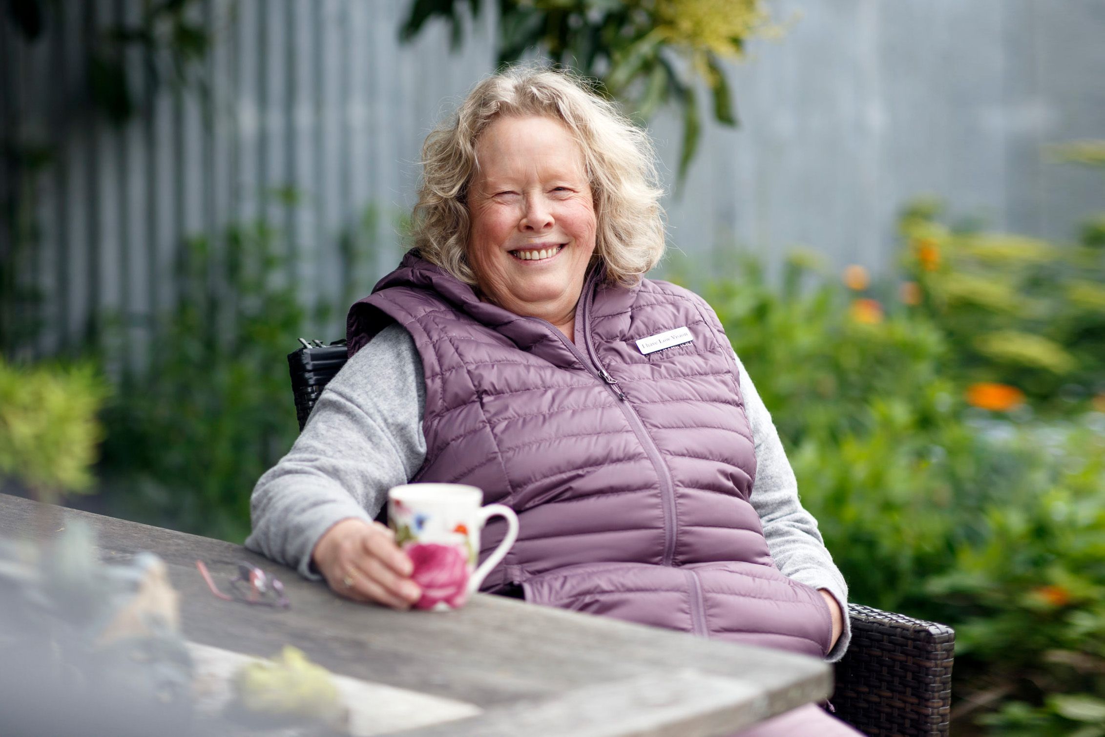 Karen wears a gilet and holds a cup of tea as she sits outside in her garden.