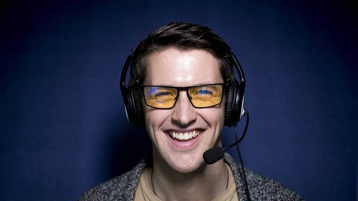 Alex Walker wears glasses and a headset, and grins at the camera in front of a dark purple background.