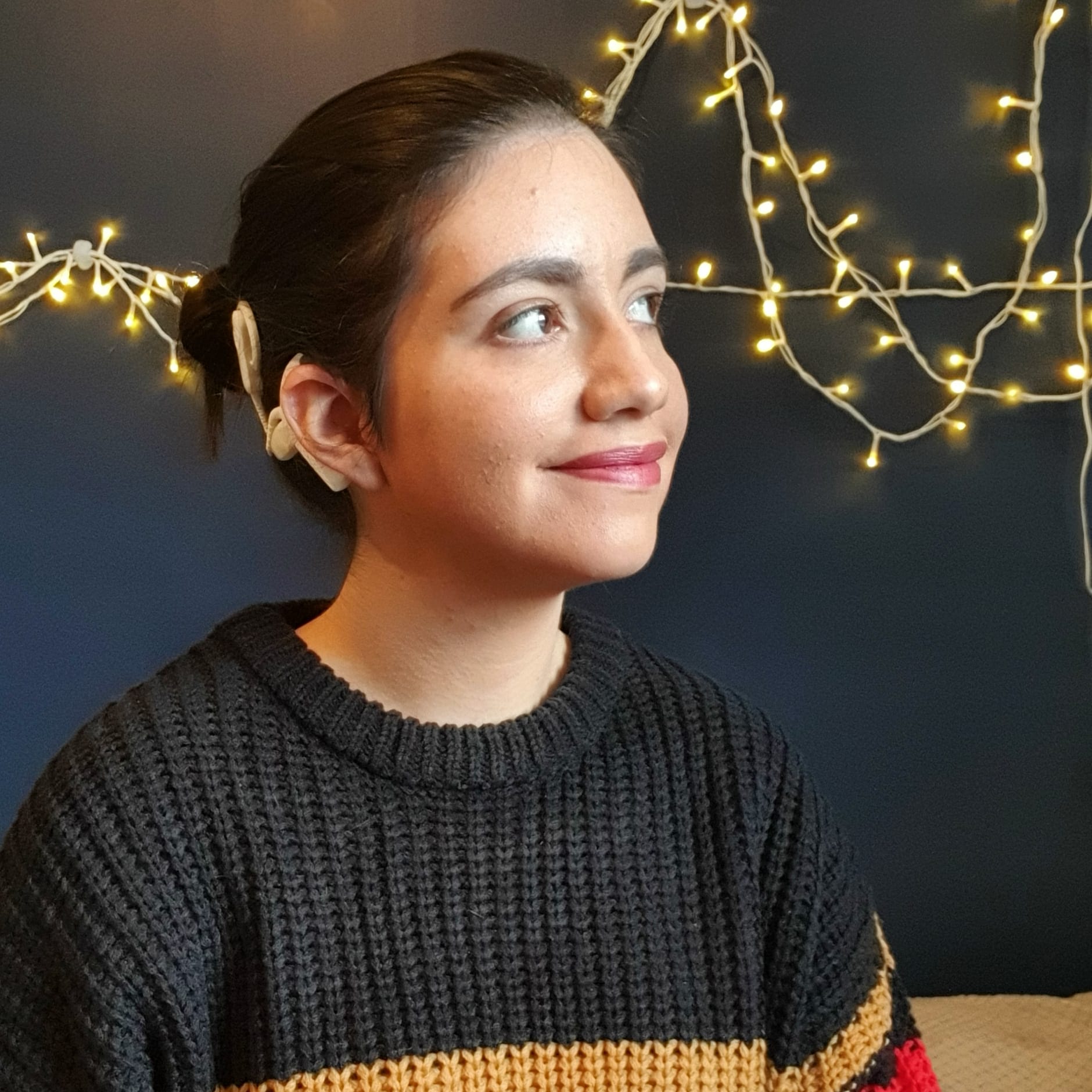 Gabriella is a young lady with dark hair, who sits in front of a blue wall with fairy lights. Her hair is tucked behind her ear, showing her cochlear implant.