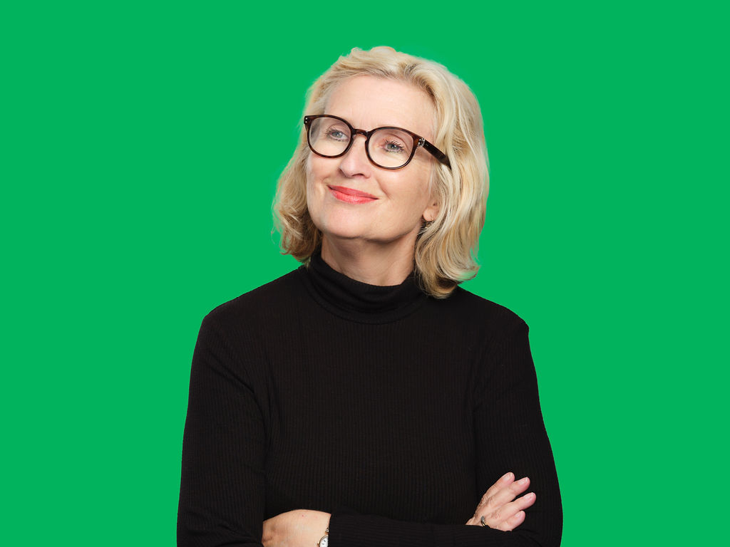 Irene has blonde hair and blue eyes, and wears a black rollneck jumper and black glasses. She smiles as she looks into the distance, with a green background behind her.