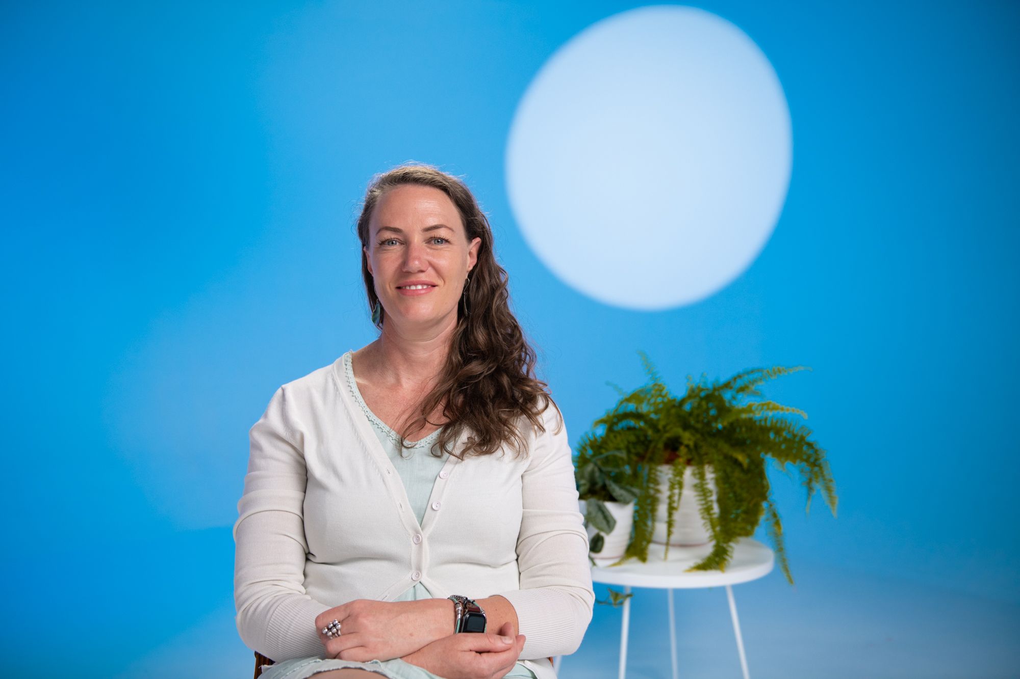 Ursula has dark blonde curly hair, green eyes, she has a relaxed smile as she looks at the camera against a blue background.