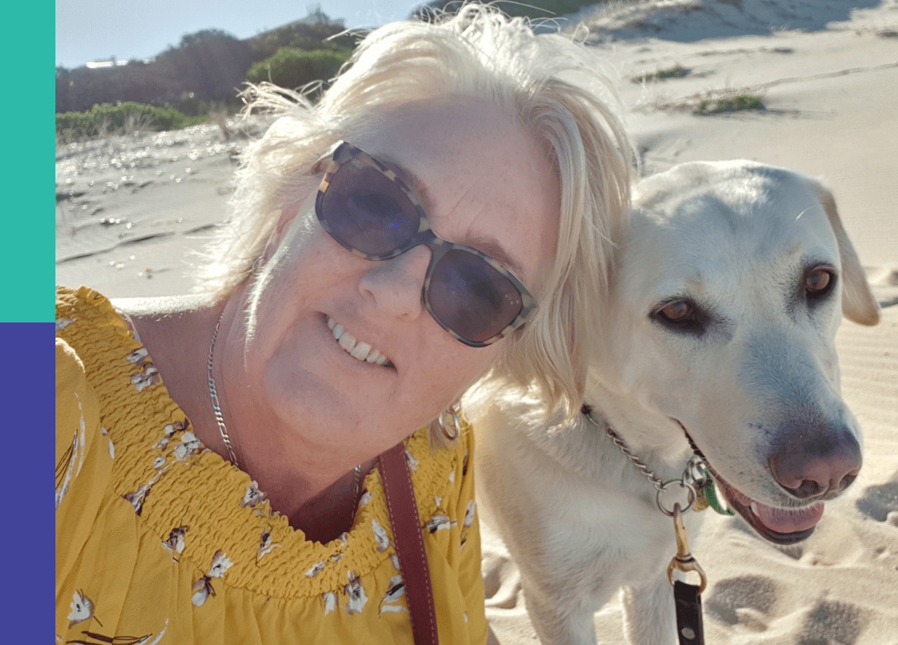 A selfie of Donna, a woman with short blonde hair and tortoise-shell glasses wearing a yellow, flower patterned blouse, sits on a beach with her guide dog, Kenzie. Kenzie, who is off harness, is looking at the camera happily.